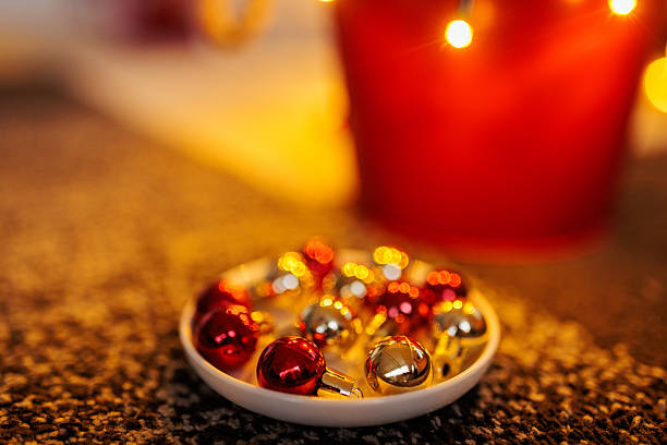 A close-up of red and gold Christmas baubles arranged in a small dish, with warm glowing lights creating a soft bokeh in the background.