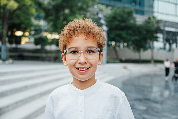 Smiling young boy with curly hair and glasses posing outdoors in a modern urban setting during the day