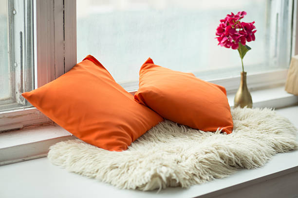 Bright orange cushions are placed on a fluffy rug near a window, with a flower vase adding a touch of elegance, enhancing the cozy home environment