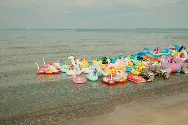Large group of colorful inflatable pool floats abandoned on a beach shoreline at the edge of the ocean, representing waste