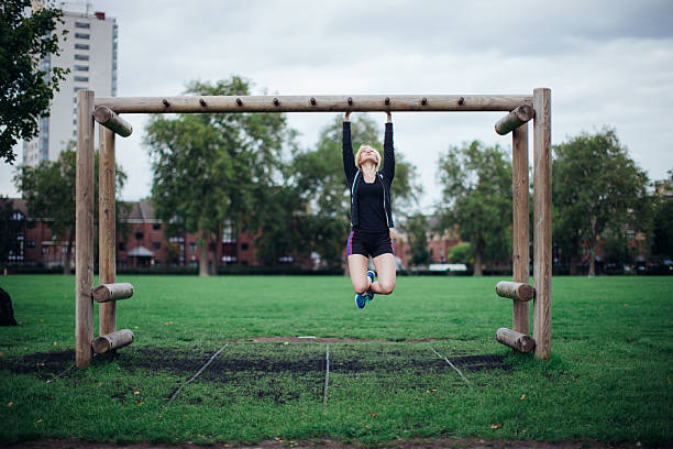A young woman hangs from a wooden exercise structure in a public park, combining playfulness with physical activity. The image highlights upper body strength, coordination, and enjoyment of movement outdoors. It represents informal fitness, freedom of movement, and using everyday environments for exercise and recreation.