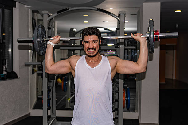 A sweaty, muscular man in a white tank top performing an intense overhead press exercise with a heavy barbell in a dimly lit fitness studio.