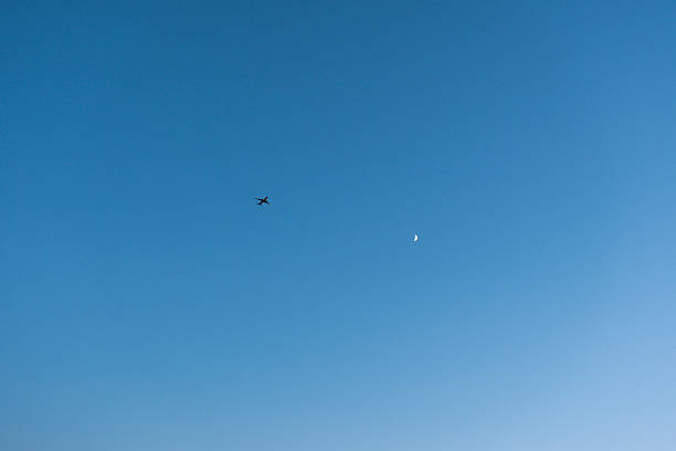 A minimalist composition featuring a small airplane and a crescent moon in a vast, clear blue sky. The wide expanse of the sky provides ample copy space, making it an ideal background for aviation, travel, or inspirational design themes. The clean blue gradient offers a professional and calm aesthetic for digital wallpapers and marketing layouts.