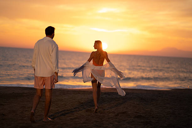 A man and woman embrace the golden hour, walking barefoot on the shore as the sun dips below the horizon, creating a serene and picturesque scene