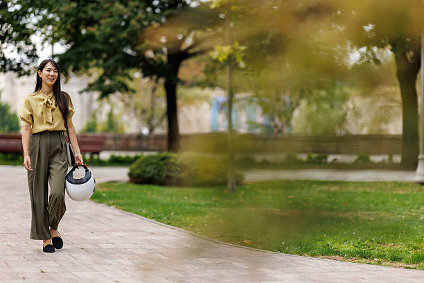 Full length shot of elegant Asian woman carrying motorcycle crash helmet while walking through green city park