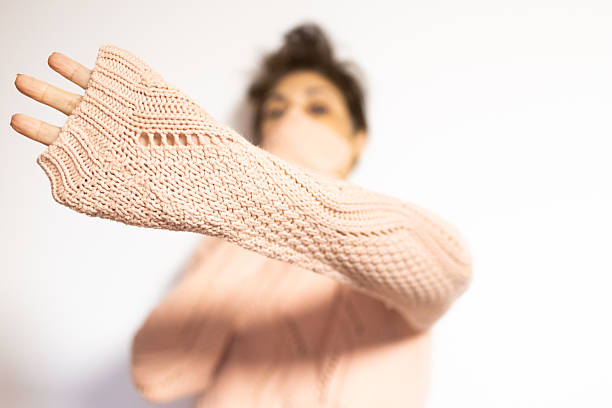 Mature middle eastern woman wearing face mask and fingerless knit sweater, extending arm in stop gesture to maintain distance and protect herself against infection, studio shot