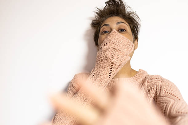 Mature middle eastern woman wearing a pink knitted sweater covering her mouth with her sleeve, showing an expression of surprise and confusion against a clean white background