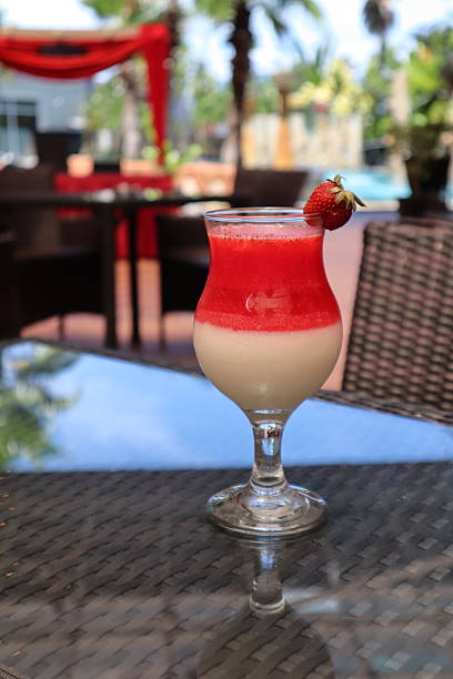 Refreshing layered drink featuring strawberry and creamy white layers, garnished with a fresh strawberry Served on a dark woven table in an outdoor setting