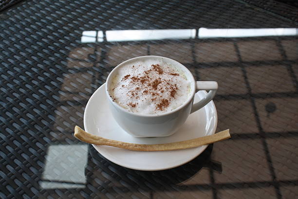Aromatic cappuccino served in a white mug, topped with frothy milk and cinnamon powder, accompanied by a long biscuit on a dark, reflective woven table