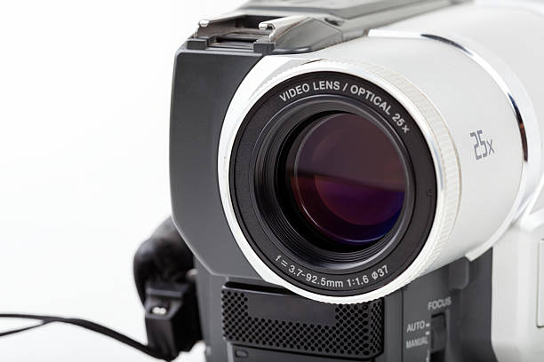 Close up of camcorder video lens with 25x optical zoom marking, front angle studio shot on white background, vintage consumer camera optics, glass reflections, focus switch details, copy space