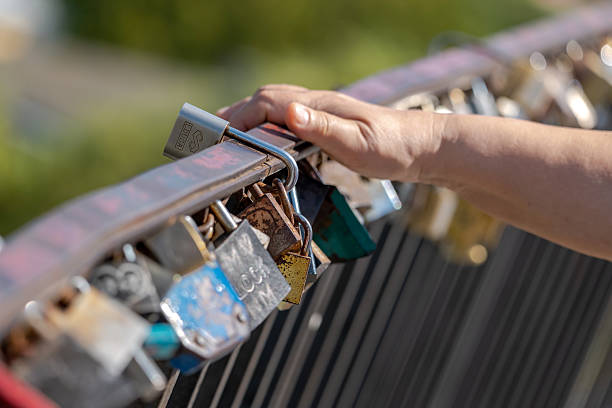 Hand touching a love lock padlock set on a bridge railing, closeup detail. Romantic tradition, eternal love symbol, couples memory concept. Metal fence with many locks, sunny day outdoors, one person