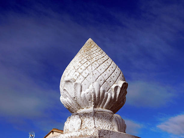 Ornate stone finial with a lotus bud design, featuring intricate carvings and a square base, set against a bright blue sky. The finial's surface appears textured and weathered, highlighting its craftsmanship and historical charm. The architectural element suggests a traditional or historic style, commonly found in palaces, temples, or significant buildings. The vivid blue sky provides a striking contrast, enhancing the finial's details and form.