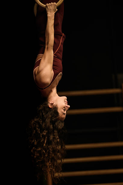 A person performs a fitness activity on rings in a dimly lit gym. The individual focuses on form and control while hanging upside down. The setting is a space dedicated to workout routines.