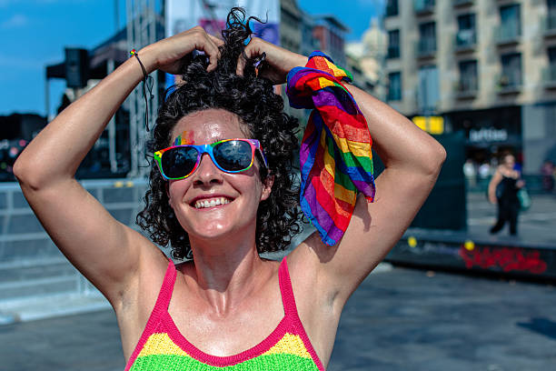 Young adult woman smiling and posing with rainbow accessories, including sunglasses, a bandana, and top, expressing joy and pride during an lgbtq+ event in a city setting