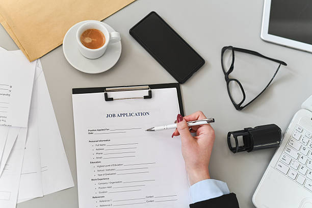 A focused professional signs a job application at a cluttered office desk, surrounded by documents, a tablet, smartphone, coffee cup, glasses, and a camera lens, conveying preparation, ambition, and administrative tasks.