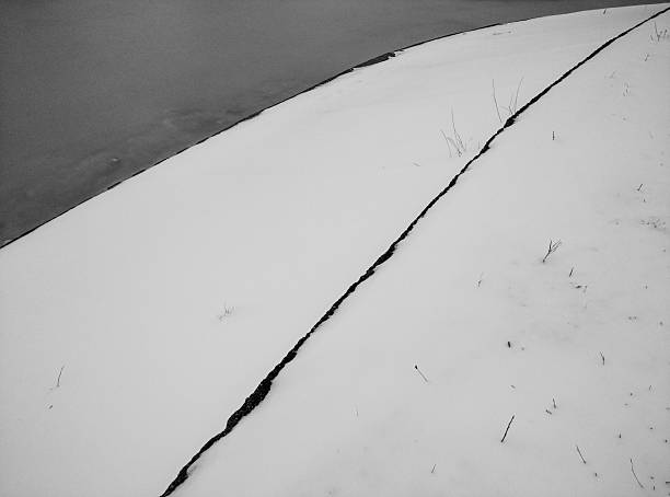 A minimalist winter scene in monochrome shows a snow-covered slope intersected by a dark diagonal line, likely a crack or seam, with water visible at the top left. The composition emphasizes contrast, stillness, and the quiet of a cold, wintry landscape.
