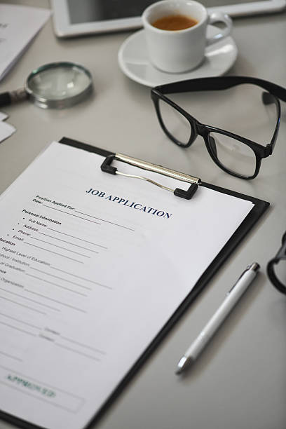 A focused businesswoman signs and reviews a job application form clipped to a clipboard in a sleek office, with glasses in the background, conveying professionalism and recruitment processes.