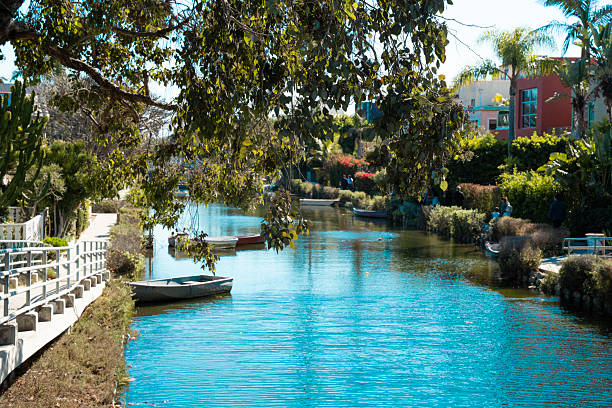 A serene waterway lined with vibrant foliage and parked boats reflects the clear blue sky, creating a picturesque urban oasis