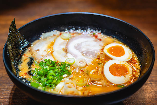 Authentic spicy Japanese ramen noodle soup with chashu pork, soft boiled eggs, and seaweed in a black ramen bowl on a wooden table.