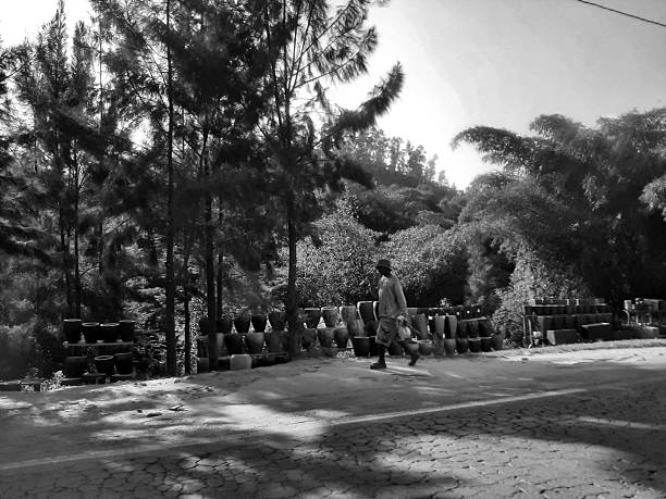 KIGALI, RWANDA - JUNE 16, 2025: A man walks along a sun-drenched road lined with various handcrafted clay pots and planters. This black and white editorial photograph highlights the traditional pottery trade and local commerce in the hilly outskirts of Rwanda.