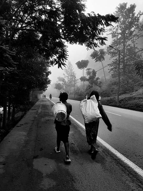 KIGALI, RWANDA - DECEMBER 28, 2024: Two people carrying loads walk along the shoulder of a paved road enveloped in thick morning fog. This black and white editorial photograph captures the atmospheric conditions and the daily commute of residents in the lush, hilly regions of Rwanda.