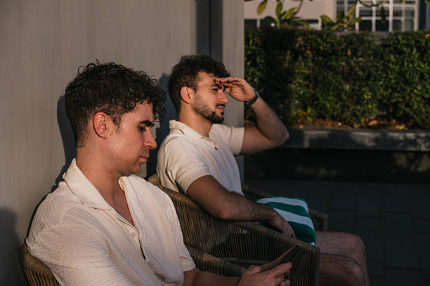 Two young adult men relaxing outdoors on a sunny day, sitting in chairs. One man uses a phone, the other shields his eyes