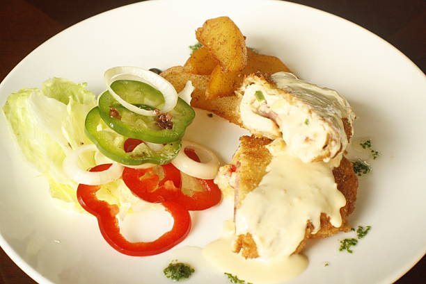 A breaded chicken roll filled with ham and peppers, covered in creamy sauce, served with roasted potatoes and a fresh vegetable salad