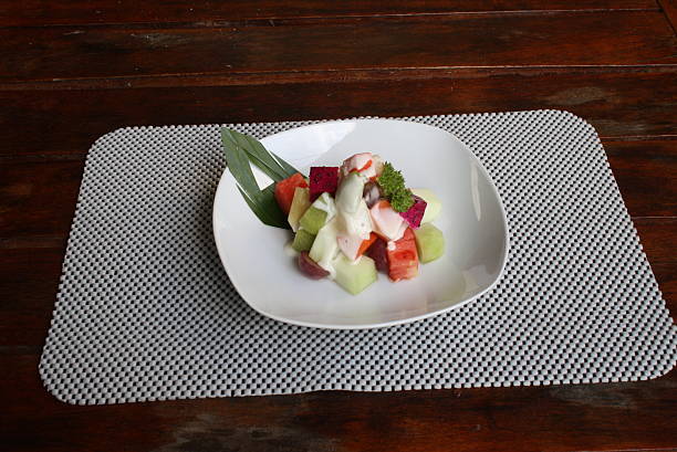 Freshly diced fruit salad featuring watermelon, dragon fruit, and melon, topped with a creamy dressing, served on a white plate and placemat