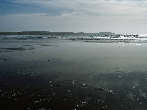 During the daytime, the expansive, wet sand beach reflects the bright sky, with gentle waves rolling in from the ocean in the distance.