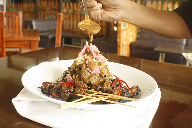 Persons hand carefully pours rich peanut sauce onto a vibrant plate of grilled skewers, fresh vegetables, and crispy crackers, served in a restaurant