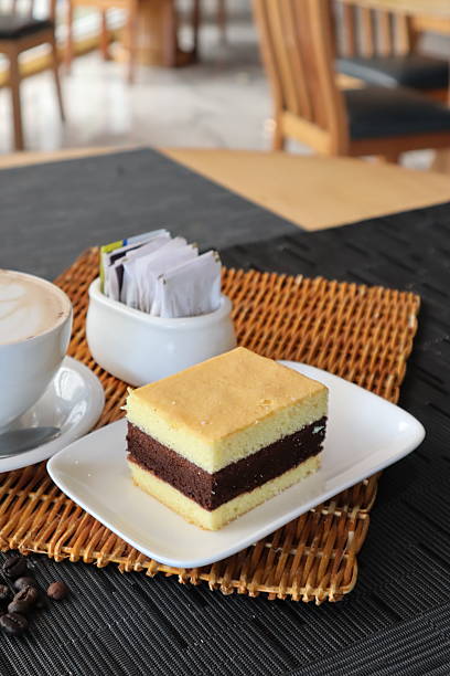 Delicious slice of layered vanilla and chocolate sponge cake served with a hot cup of coffee and sugar packets on a textured placemat, perfect for a break