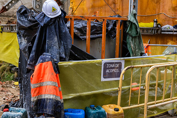 A still life of labor: a mud-stained orange reflective jacket and a white hard hat rest on equipment at a construction site. Surrounded by blue chemical jerrycans and dirty tarps, these personal protective equipment (PPE) items symbolize the human element, labor breaks, or the conclusion of a shift in the building industry.