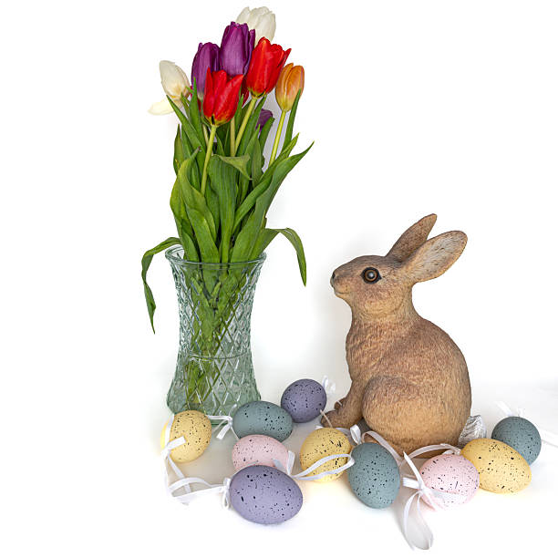 Easter bunny surrounded by Easter eggs looking at multi-coloured tulips in a crystal glass vase.