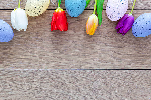 Five multi-coloured Easter eggs and four tulips at the top edge of a brown table with planks in a horizontal line.
