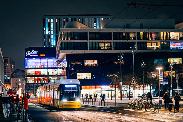 Berlin, Germany – January 31, 2026: A tram travels through a city street at night as pedestrians move along the sidewalks near the CineStar Cubix cinema complex at Alexanderplatz. Snow and icy patches are visible on the roadway and tram tracks, creating slippery conditions and traffic safety risks during winter weather. The scene relates to public transportation service disruptions and potential delays during strike action.