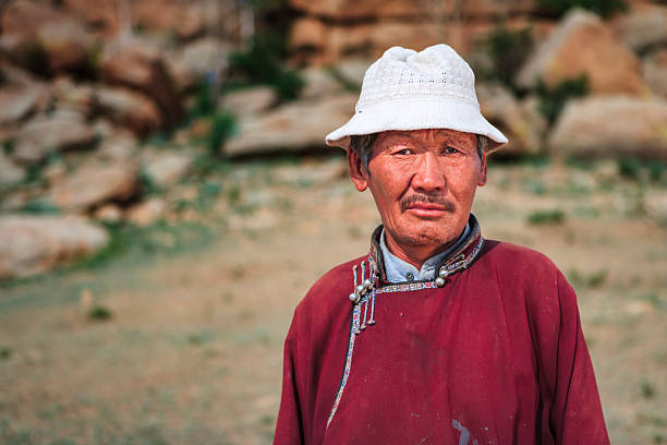 A weathered older man wearing a deep red traditional robe with decorative trim and metal fastenings stands outdoors. He has a white knit bucket hat, a mustache, and a sun-tanned, lined face, looking directly at the camera with a serious, contemplative expression. The background is softly blurred, showing rocky outcrops and sparse grass that suggest a rugged, open landscape.