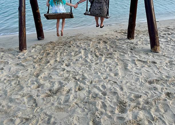 Wide view of a mother and child enjoying a rustic wooden swing installed directly on a sandy tropical beach. Ocean water and textured sand create a serene summer vacation atmosphere, symbolizing family bonding, freedom, and carefree coastal living.