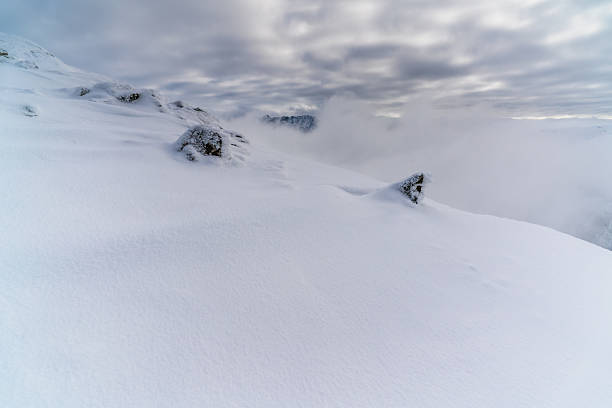 Snow-covered mountain ridge with smooth wind-shaped snow and exposed rocks, while dense clouds and fog drift below, partially hiding distant peaks and creating a stark, isolated winter atmosphere.