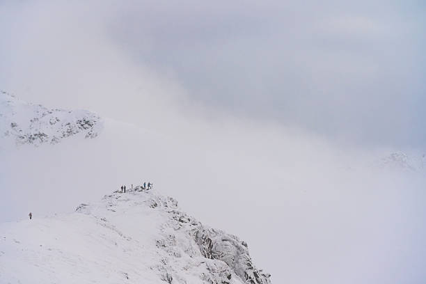 A narrow, snow-covered ridge rises above a sea of fog, with tiny figures emphasizing scale. Soft winter light and minimal composition create a calm, alpine atmosphere.