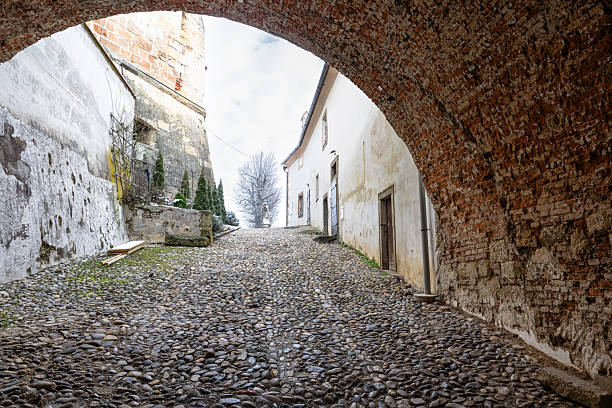A cobblestone street runs beneath a weathered brick archway, flanked by aged walls and doors. Light snowfall and muted tones evoke a quiet, timeless village atmosphere.