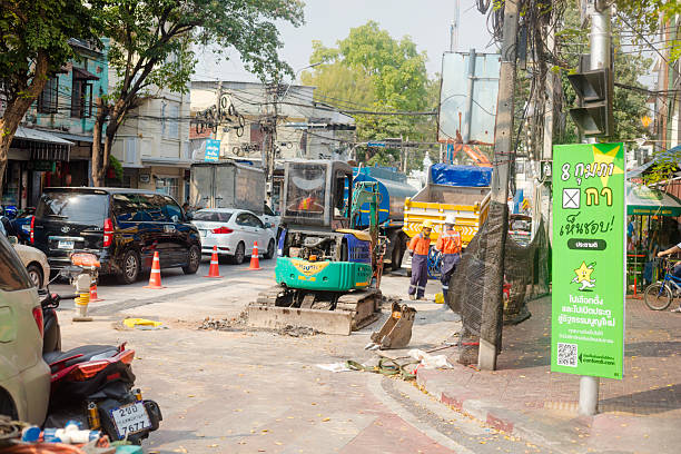 Small street construction site in Bangkok Phra Nakhon at small intersection of streets Bamrung Mueang Road and Maha Chai Road. In background are old buildings and rowhouses. Two thai construction workers are stranding near a small digger. At right side is a billboard for actual election. Traffic is queued in Maha Chai Road.