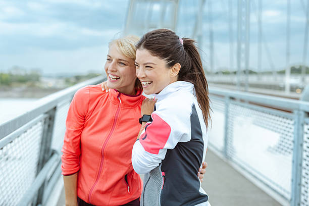 Two mid‑adult women jog side by side on a bridge in the city, smiling and enjoying a healthy lifestyle together.