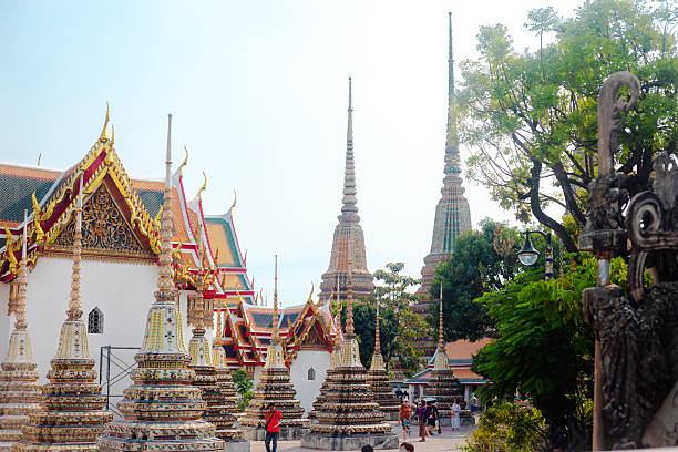 View into ground of Wat Pho monastery with many temples ans stupas colorful decorated in thai art and mosaic. View over wall from Thai Wang Road in Phra Nakhon at afternoon. A few people are visible in bottom area.