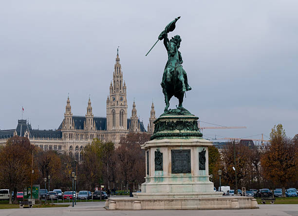 This picture shows the Vienna City Hall (Rathaus) silhouetted against the bright afternoon sun. The central tower with its clock and spire rises prominently, flanked by smaller towers with intricate Gothic Revival detailing.