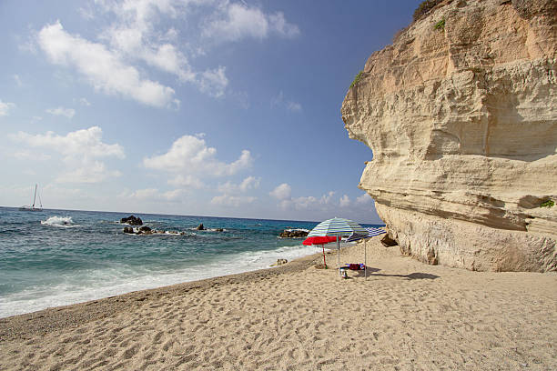 Santa Domenica di Ricadi, Calabria, Italy: scenic view of Riaci Bay beach. A massive, overhanging limestone cliff dominates the right side of the image, sheltering colorful sun umbrellas on the sandy shore. To the left, turquoise waves crash against rocks in the sea, with a sailboat visible on the horizon under a cloudy blue sky.