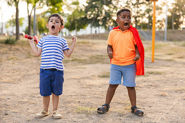 Young boys playing pretend superhero outdoors, one wearing a red cape and shouting, capturing the joy of childhood imagination