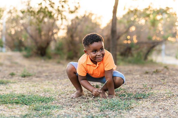 Happy black boy squatting on green grass, smiling and exploring nature with a stick on a sunny summer day, carefree curiosity