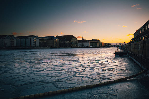 Ice covers Copenhagen Harbor in Denmark as an unusual winter cold snap freezes sections of the city's normally navigable waterways on February 3, 2026. The frozen harbor shows buildings along the waterfront silhouetted against the sunset sky, with thick ice formations visible across the water surface. The extreme winter weather event has created unusual conditions in the Danish capital's harbor district.
