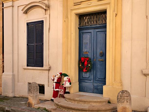 Traditional Maltese architecture with blue arched double door, dark shuttered window, anda playful Santa Claus and snowman display for Christmas. The sunny scene on alimestone facade captures festive Mediterranean street charm.
