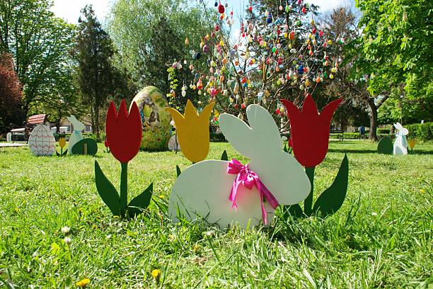Colorful Easter decorations in a city park during springtime. A large hand-painted Easter egg and an egg tree stands among decorative easter bunny figures and tulip ornaments on green grass, surrounded by blooming trees and fresh foliage. Easter egg hunt. Festive outdoor scene celebrating Easter holiday, joy, renewal, and seasonal tradition. Ideal for themes of spring, family celebration, childhood, holidays, urban nature, and cheerful public events.
